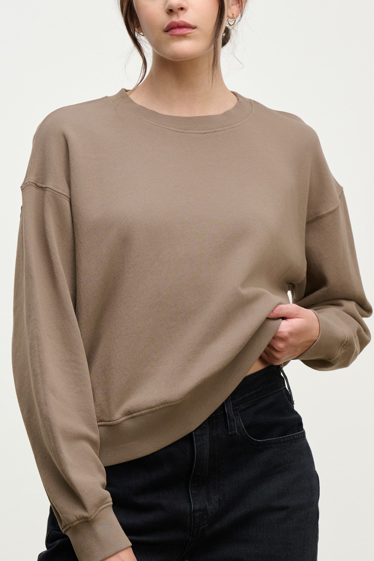 Wearing the relaxed silhouette of the YOKO SWEATSHIRT by Velvet by Graham & Spencer in soft cotton fleece, a person stands against a plain background, holding the hem of their brown long-sleeve top paired with black jeans.-40328390836417
