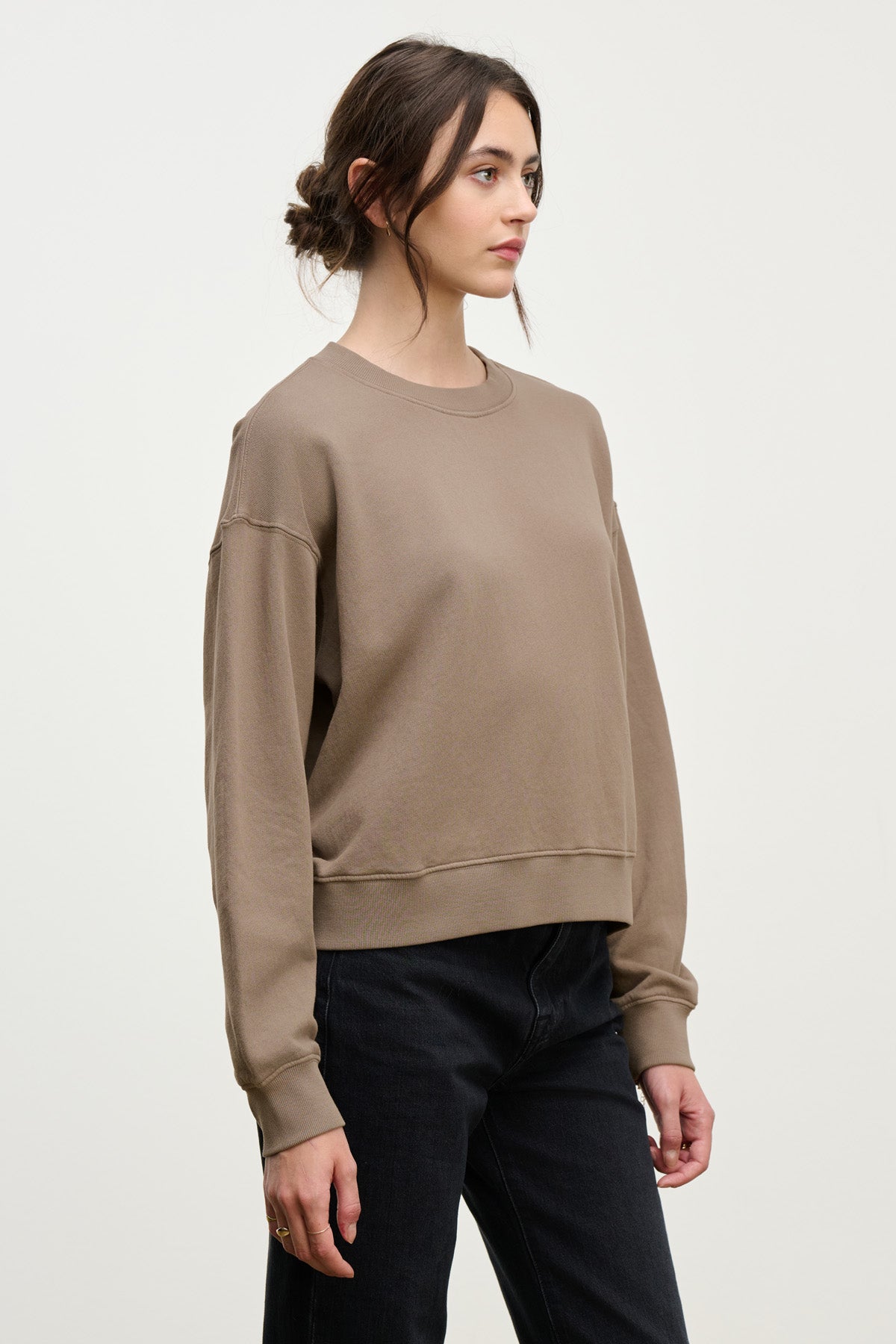 A woman with brown hair in a low bun wears the YOKO SWEATSHIRT by Velvet by Graham & Spencer, featuring a relaxed silhouette, paired with black jeans and standing against a plain light background.-40328390869185