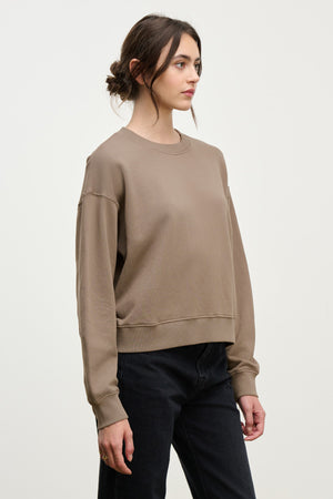 A woman with brown hair in a low bun wears the YOKO SWEATSHIRT by Velvet by Graham & Spencer, featuring a relaxed silhouette, paired with black jeans and standing against a plain light background.