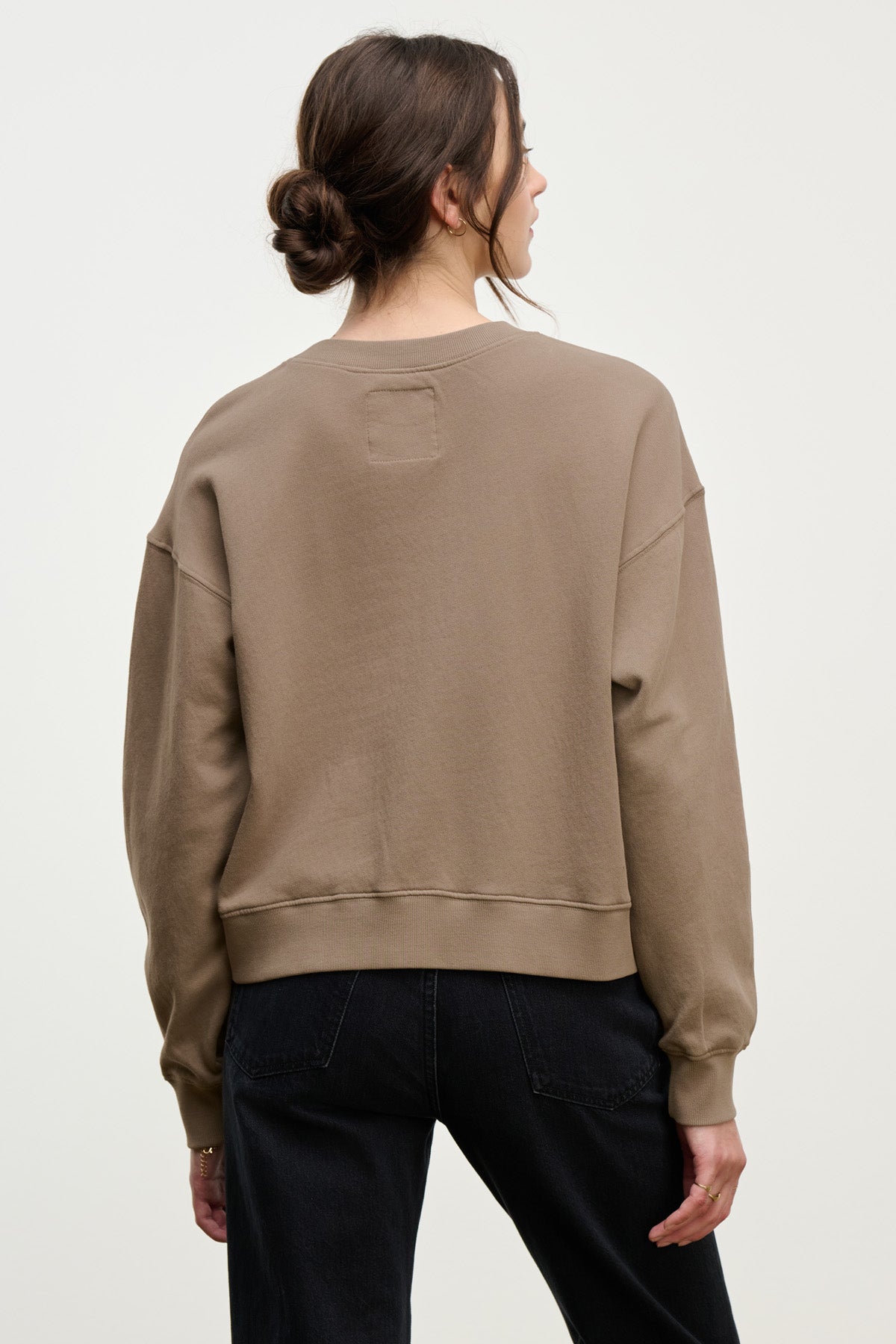 A person with brown hair in a low bun wears the taupe YOKO SWEATSHIRT by Velvet by Graham & Spencer, featuring a relaxed silhouette, paired with black pants, standing with their back to the camera against a plain light background.-40328390901953