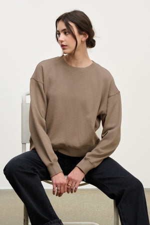 A woman sits on a white chair in a relaxed pose, wearing the YOKO SWEATSHIRT by Velvet by Graham & Spencer in soft cotton fleece and black pants, gazing to her left against a plain background.