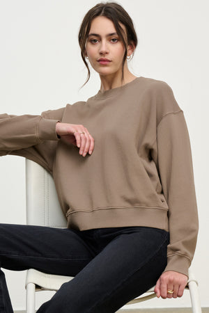 A woman with dark hair in a low bun wears the Velvet by Graham & Spencer YOKO SWEATSHIRT in taupe with black jeans, creating a relaxed look as she sits on a white chair against a plain background.