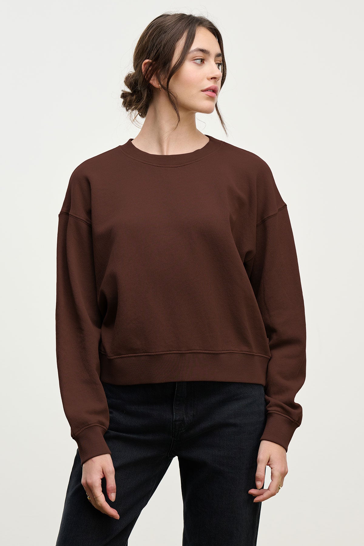 A woman stands against a plain background, wearing the Velvet by Graham & Spencer YOKO SWEATSHIRT in brown cotton fleece with a relaxed fit, paired with black pants. Her hair is tied back as she looks to the side.-40416929087681