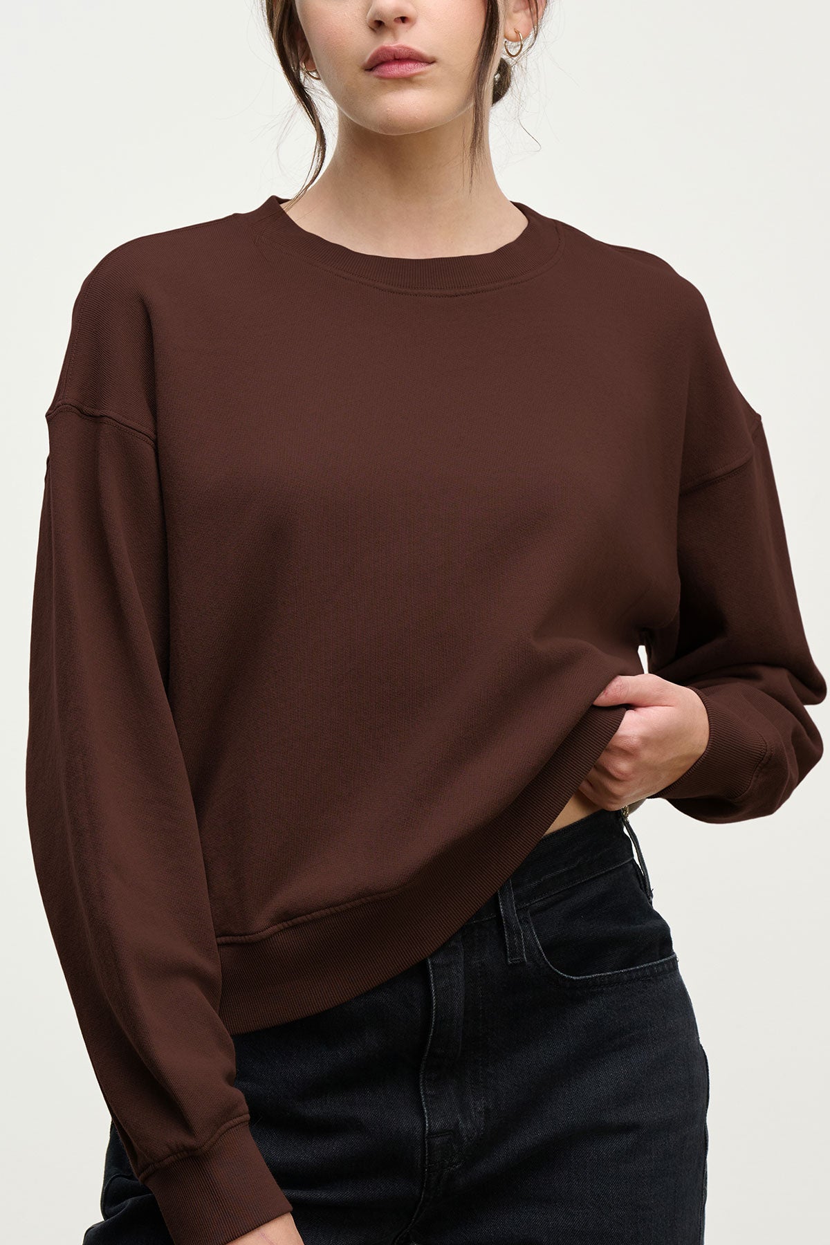 A woman wears the Velvet by Graham & Spencer YOKO Sweatshirt in plain brown cotton fleece with a relaxed fit and black jeans, holding up the hem with one hand against a neutral background.