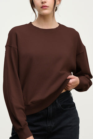 A woman wears the Velvet by Graham & Spencer YOKO Sweatshirt in plain brown cotton fleece with a relaxed fit and black jeans, holding up the hem with one hand against a neutral background.