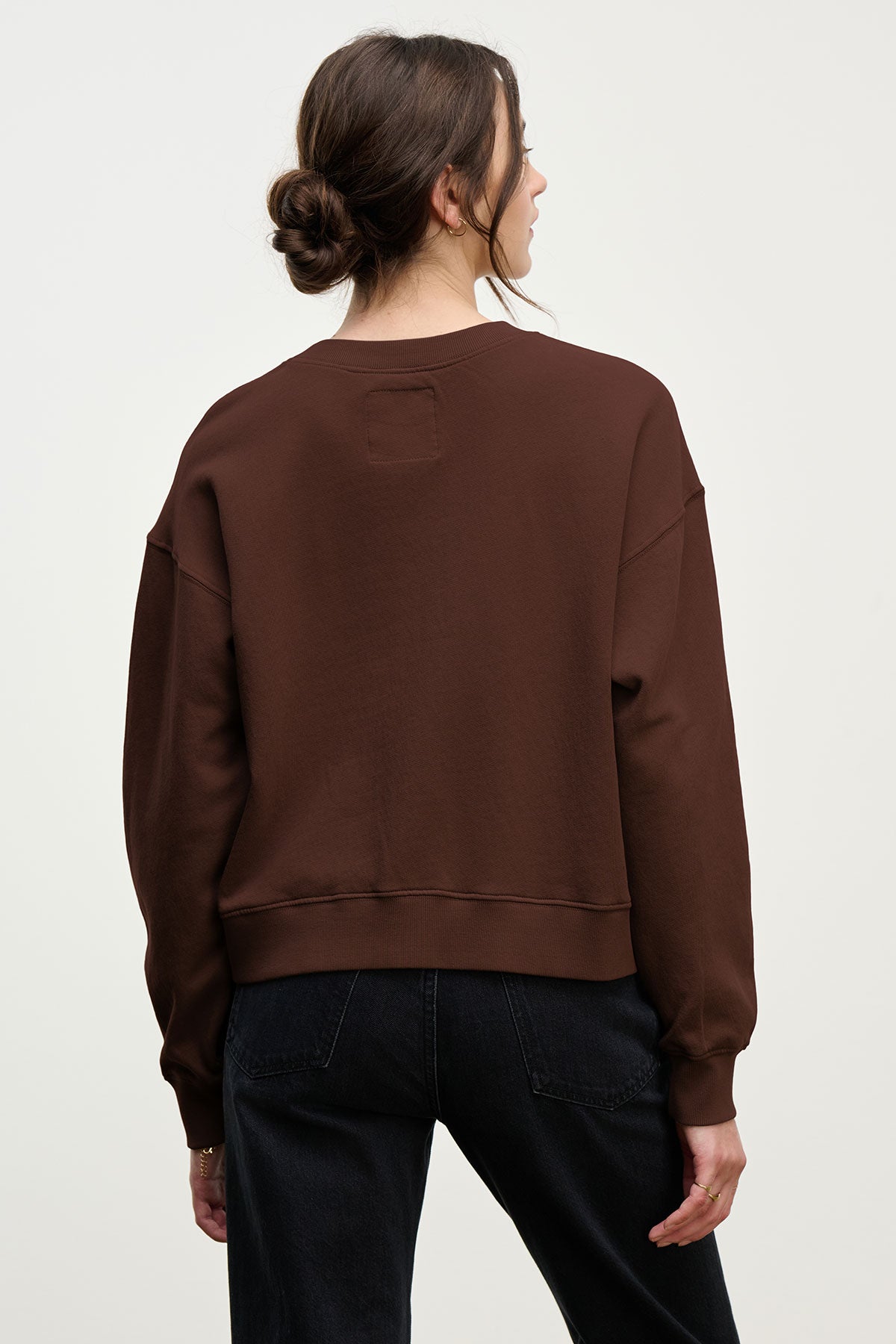 A woman with brown hair in a low bun wears a relaxed-fit YOKO SWEATSHIRT by Velvet by Graham & Spencer and black jeans, standing with her back to the camera against a plain background.