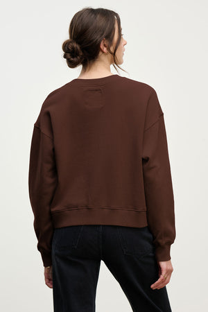 A woman with brown hair in a low bun wears a relaxed-fit YOKO SWEATSHIRT by Velvet by Graham & Spencer and black jeans, standing with her back to the camera against a plain background.