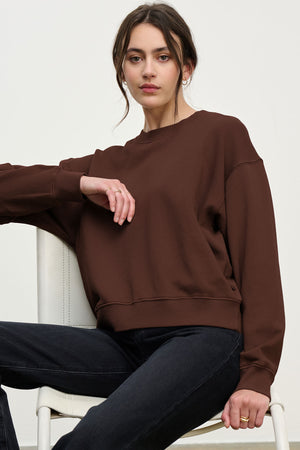 A woman wearing the Velvet by Graham & Spencer YOKO SWEATSHIRT in a relaxed silhouette with black jeans sits on a white chair against a plain background, looking directly at the camera.
