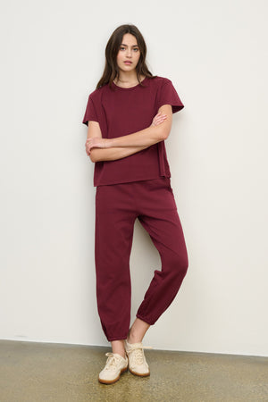 A woman stands against a plain wall wearing a relaxed fit burgundy short-sleeve top and Velvet by Graham & Spencer ZARA SWEATPANT joggers, paired with beige sneakers.