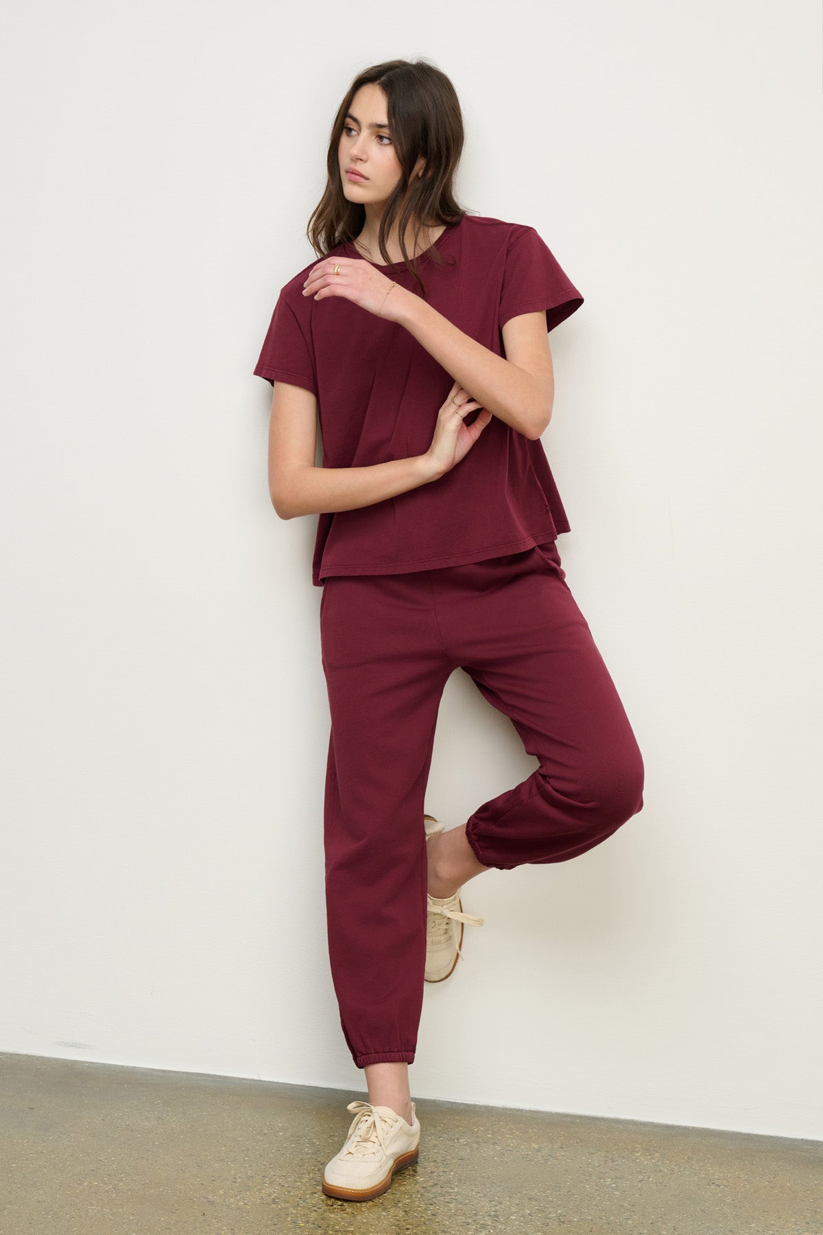 A woman wearing a matching maroon short-sleeve top and Velvet by Graham & Spencer ZARA SWEATPANT leans against a white wall with one leg bent and arms crossed.
