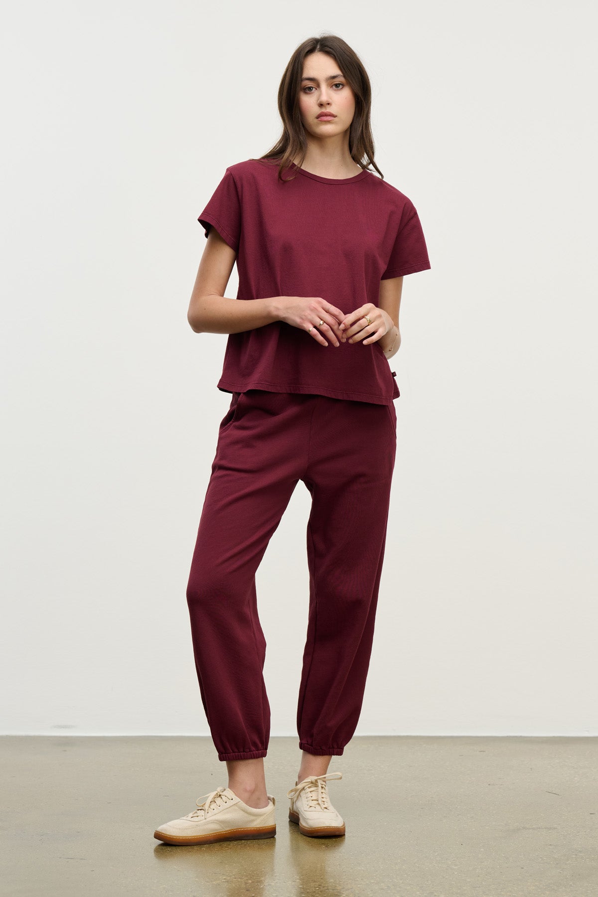 A woman stands against a plain background wearing a relaxed fit burgundy t-shirt and Velvet by Graham & Spencer ZARA SWEATPANT set, paired with beige sneakers.-40328398700737