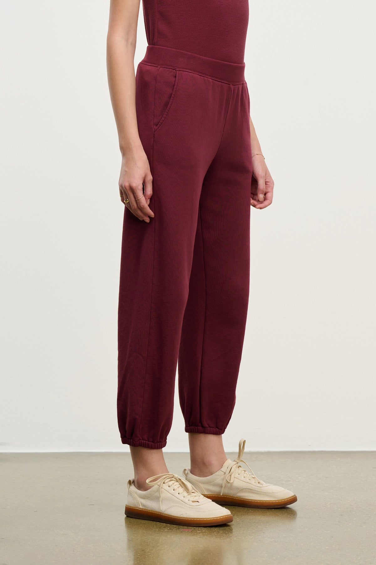 A person wearing relaxed fit ZARA SWEATPANT joggers by Velvet by Graham & Spencer in maroon with beige sneakers stands against a plain off-white wall.-40328398766273