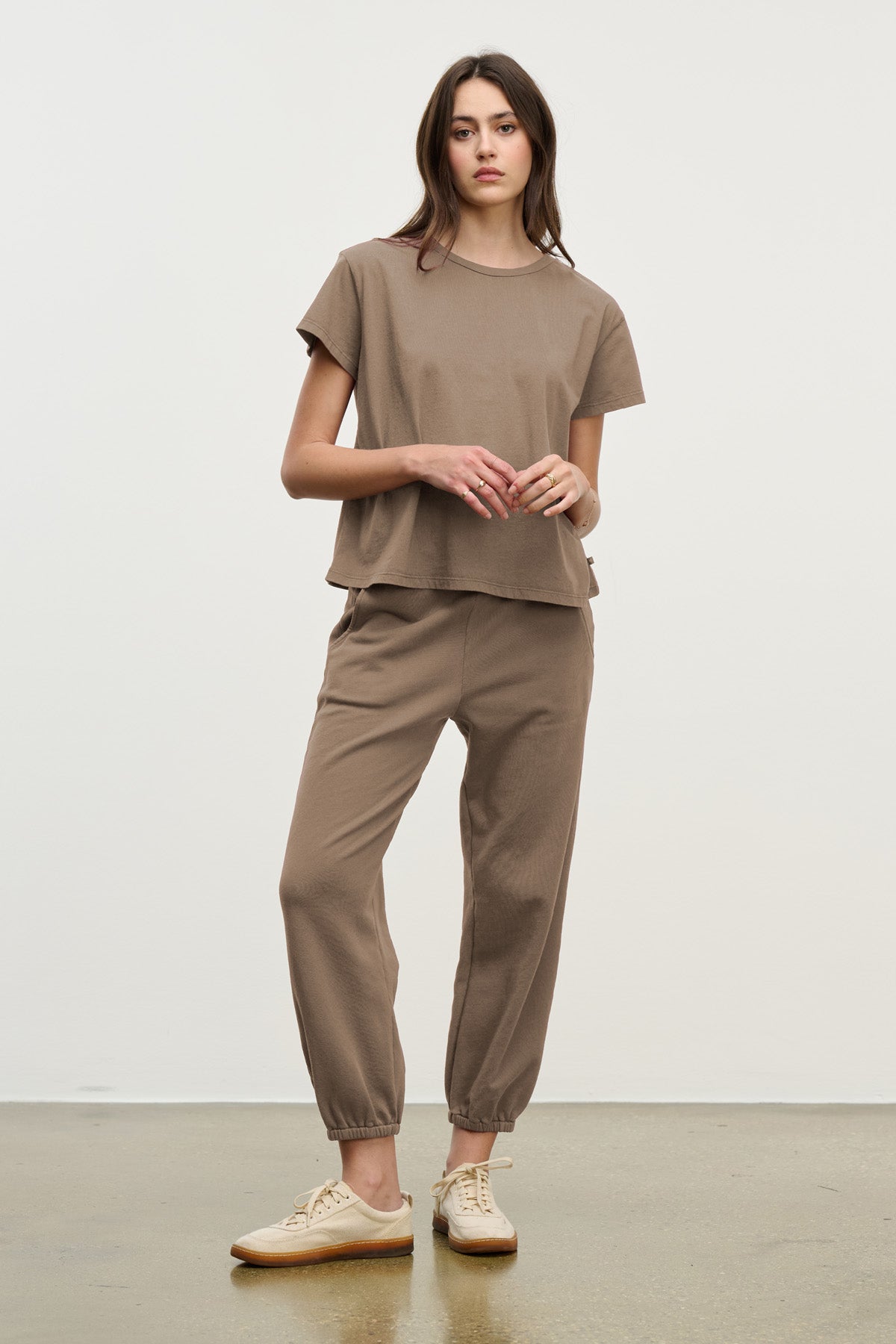 A person stands against a plain background wearing a relaxed fit brown T-shirt, ZARA SWEATPANT joggers by Velvet by Graham & Spencer crafted from cotton fleece, and beige sneakers.-40328398864577