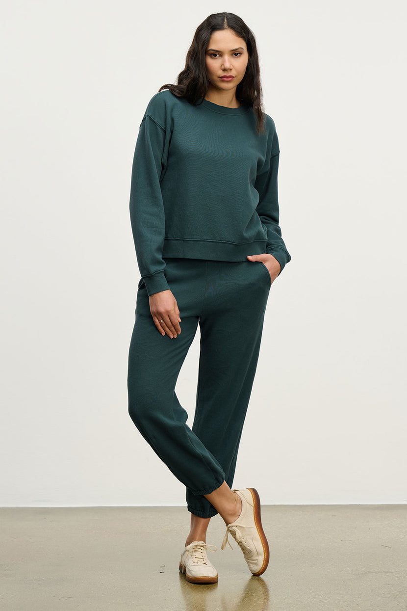A woman stands against a plain background wearing Velvet by Jenny Graham’s ZUMA SWEATPANT in dark green organic cotton with a matching sweatshirt and beige sneakers.