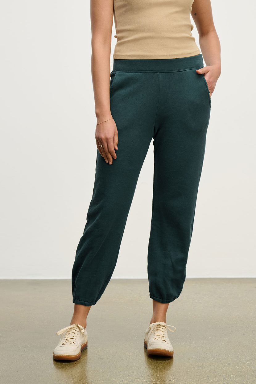 A person in a beige sleeveless top, Velvet by Jenny Graham's ZUMA SWEATPANT in dark green organic cotton with pockets, and beige sneakers stands on a polished floor.