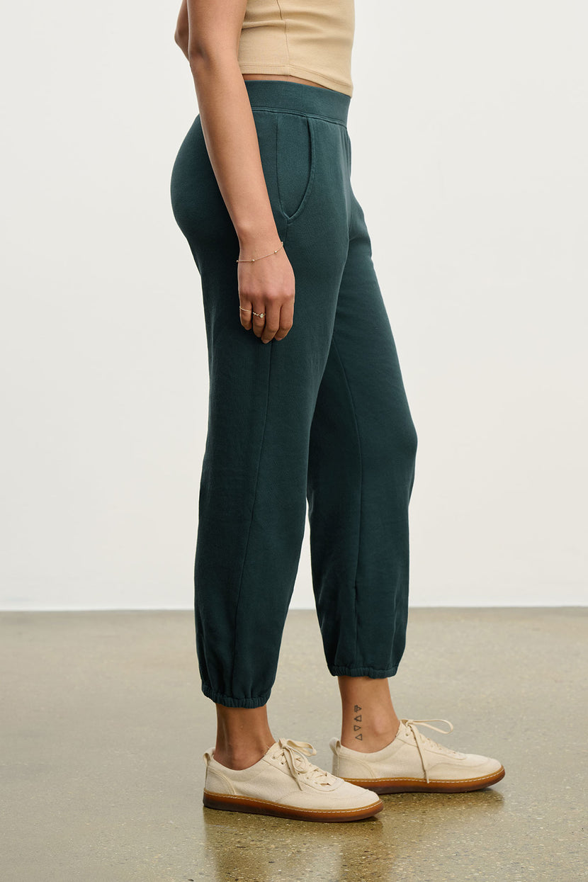 Someone stands on a polished floor, photographed from the side, wearing Velvet by Jenny Graham's ZUMA SWEATPANT in dark green organic cotton, paired with a beige top and beige lace-up shoes.