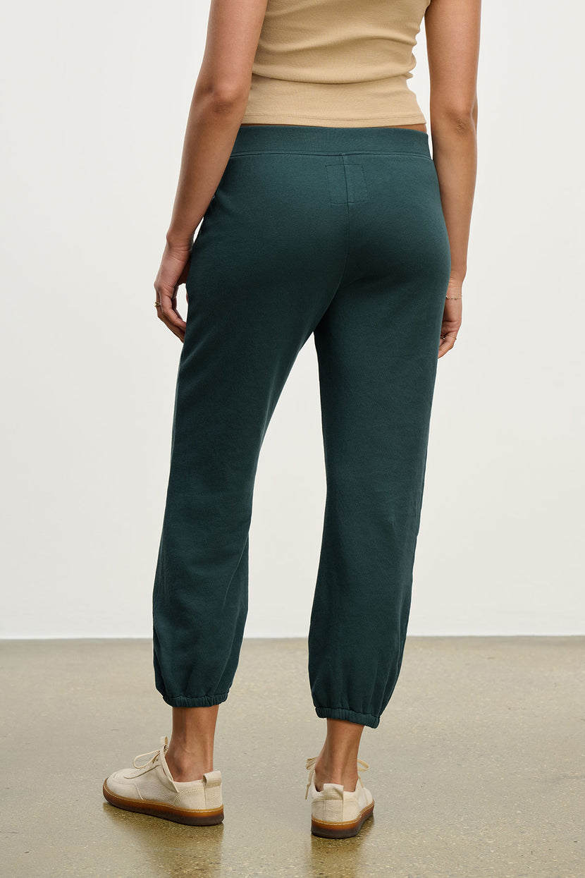 A person stands facing away, wearing Velvet by Jenny Graham's ZUMA SWEATPANT in dark green with a beige sleeveless top and beige sneakers, on a polished floor against a plain white background.