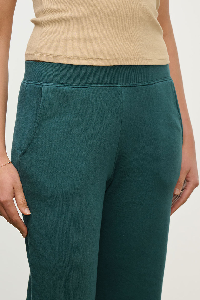 A person wearing a beige sleeveless top and Velvet by Jenny Graham’s ZUMA SWEATPANT in dark green organic cotton, with hands resting at their sides.