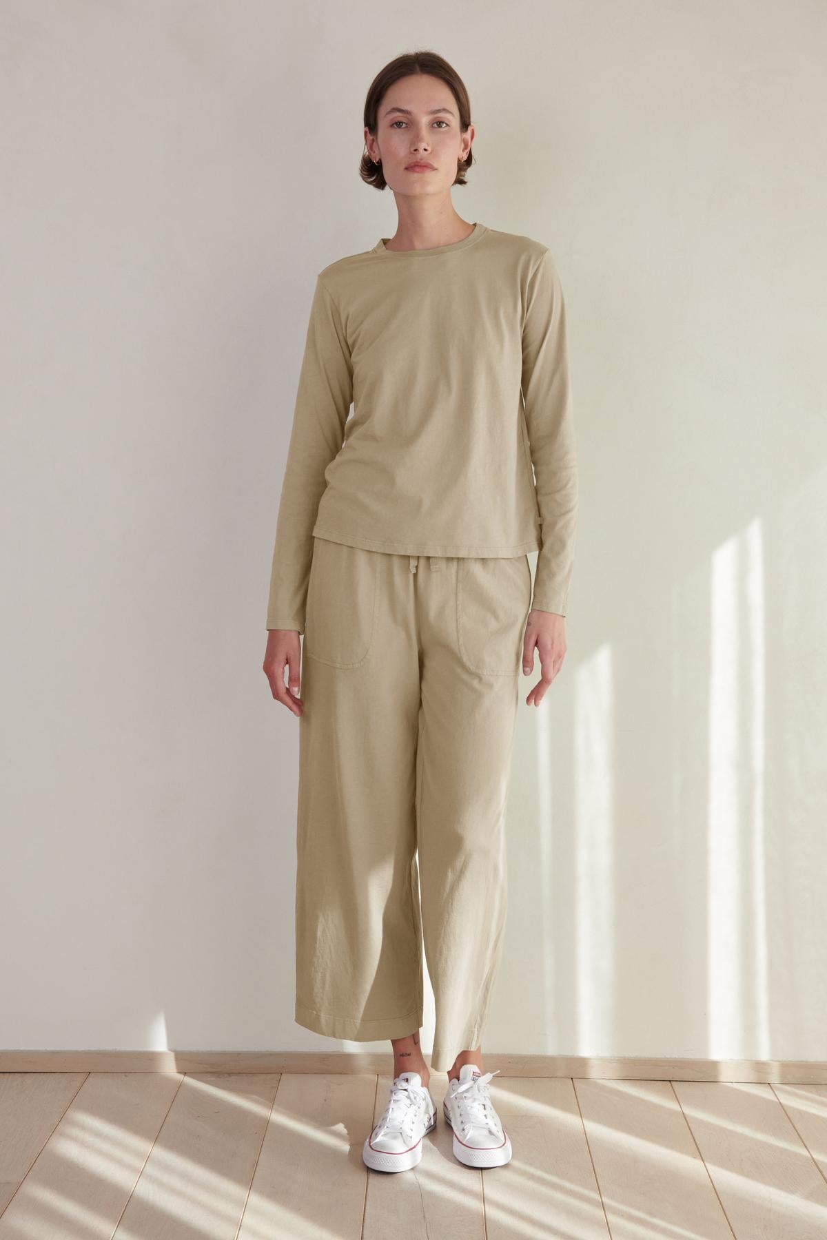 Woman standing in a room wearing an organic cotton beige long-sleeve top and matching Velvet by Jenny Graham PISMO PANT with white sneakers.-36463776432321