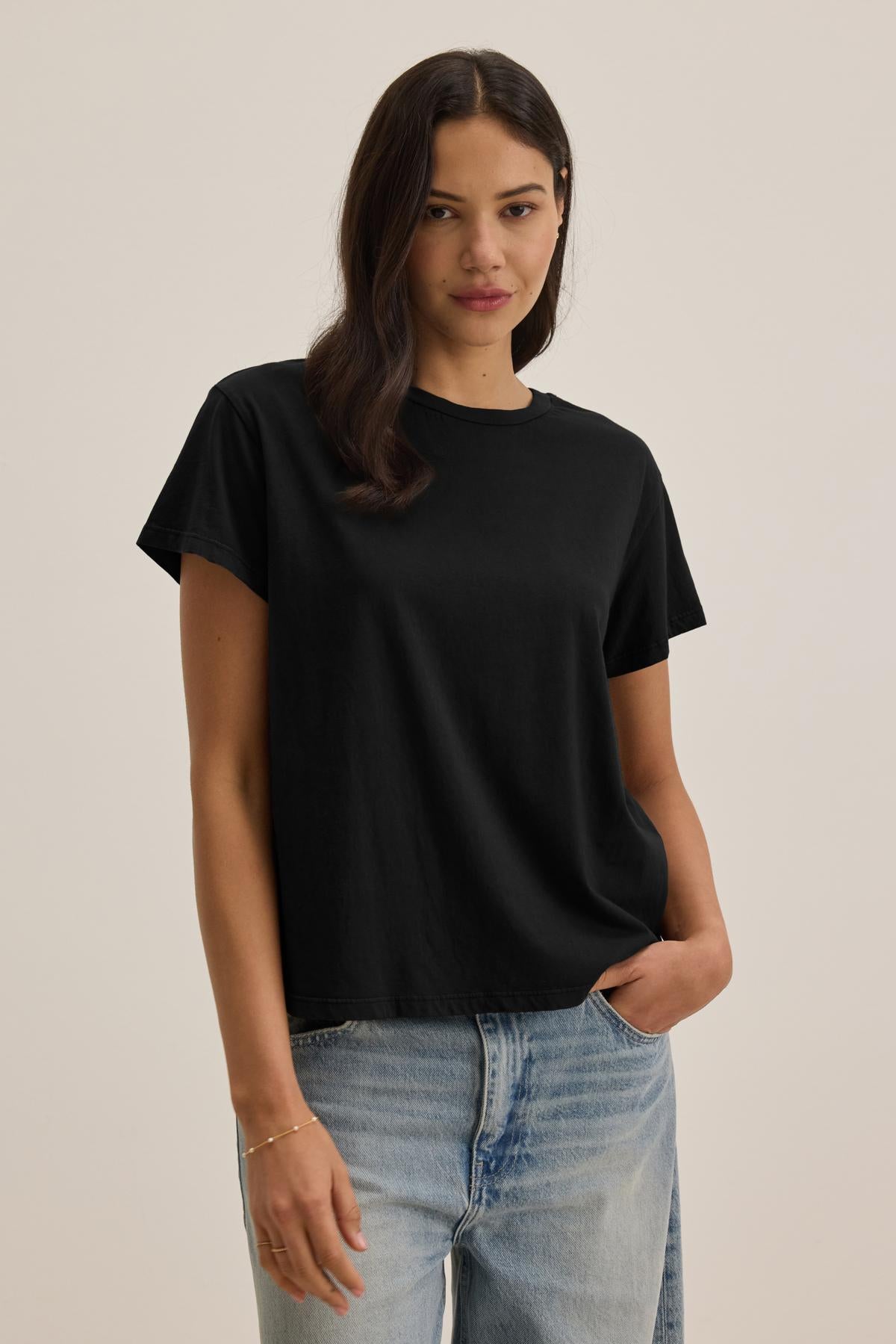   A woman with long dark hair wears the TOPANGA TEE from Velvet by Jenny Graham, a relaxed-fit classic black t-shirt, paired with organic cotton jeans. She strikes a pose with one hand in her pocket against a simple, eco-friendly backdrop. 