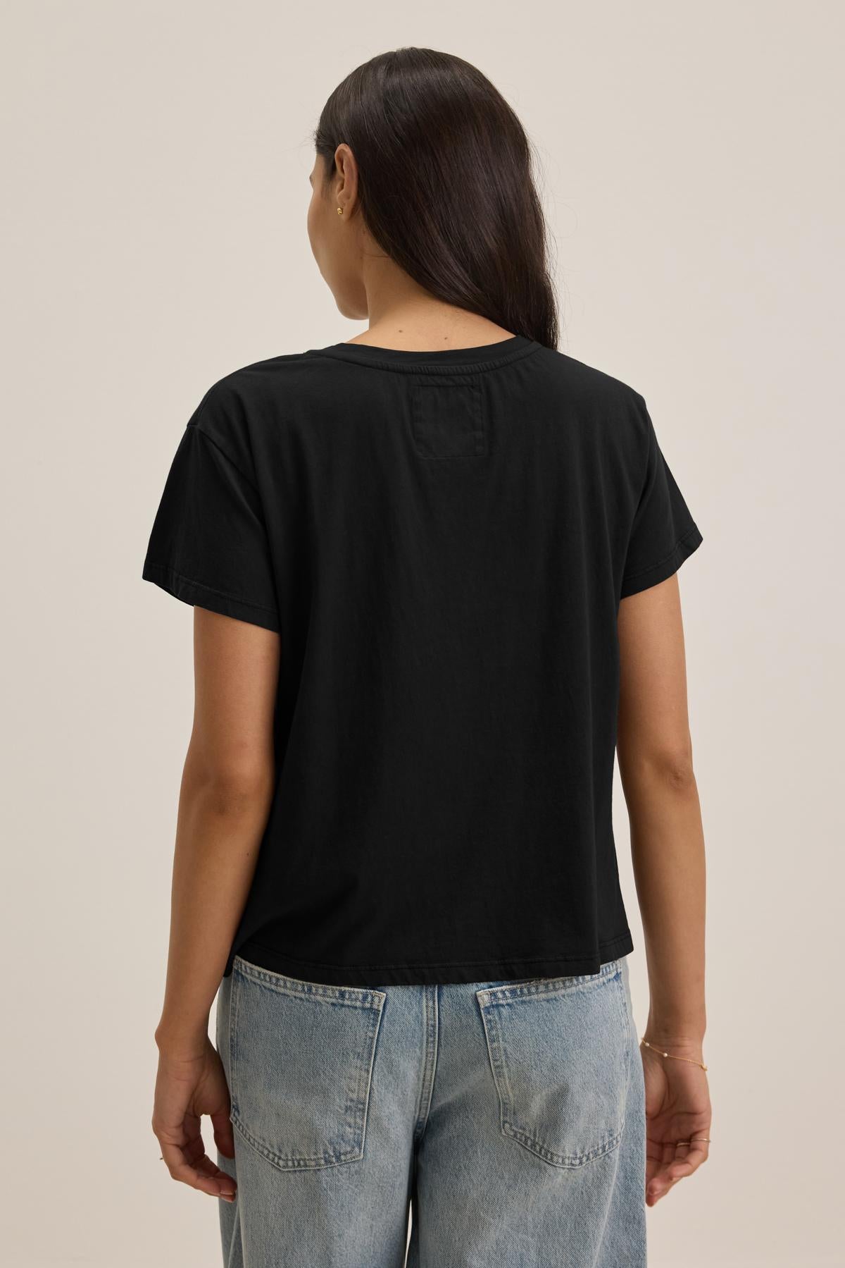   A person with long dark hair is shown from the back, wearing the Velvet by Jenny Graham TOPANGA TEE, a black short-sleeve t-shirt made of organic cotton, paired with light blue, relaxed-fit classic jeans. 