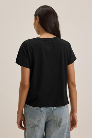 A person with long dark hair is shown from the back, wearing the Velvet by Jenny Graham TOPANGA TEE, a black short-sleeve t-shirt made of organic cotton, paired with light blue, relaxed-fit classic jeans.