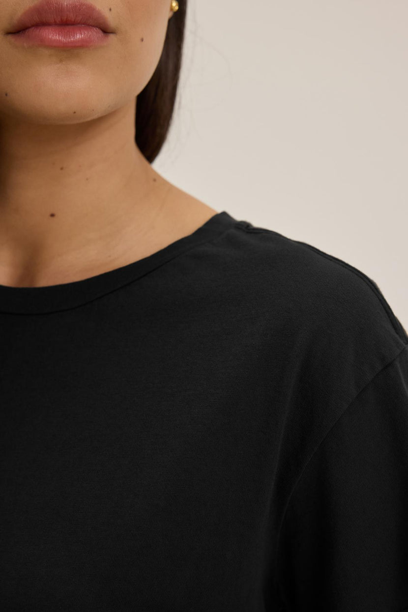 Close-up of a person with long brown hair, wearing a relaxed-fit classic black TOPANGA TEE made of organic cotton by Velvet by Jenny Graham. Only the lower half of the face and shoulder are visible.