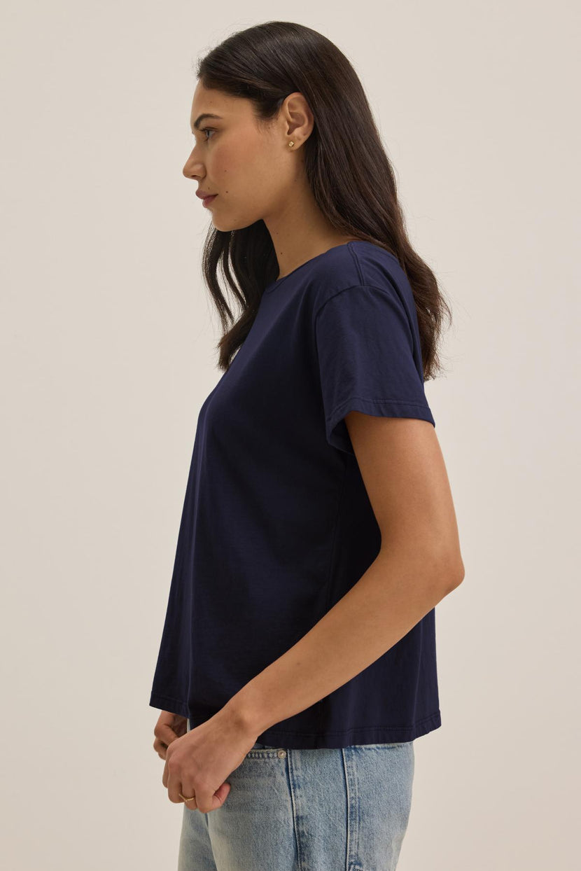 Side profile of a person with long, dark hair wearing the navy blue TOPANGA TEE by Velvet by Jenny Graham and light blue jeans, standing against a plain background.