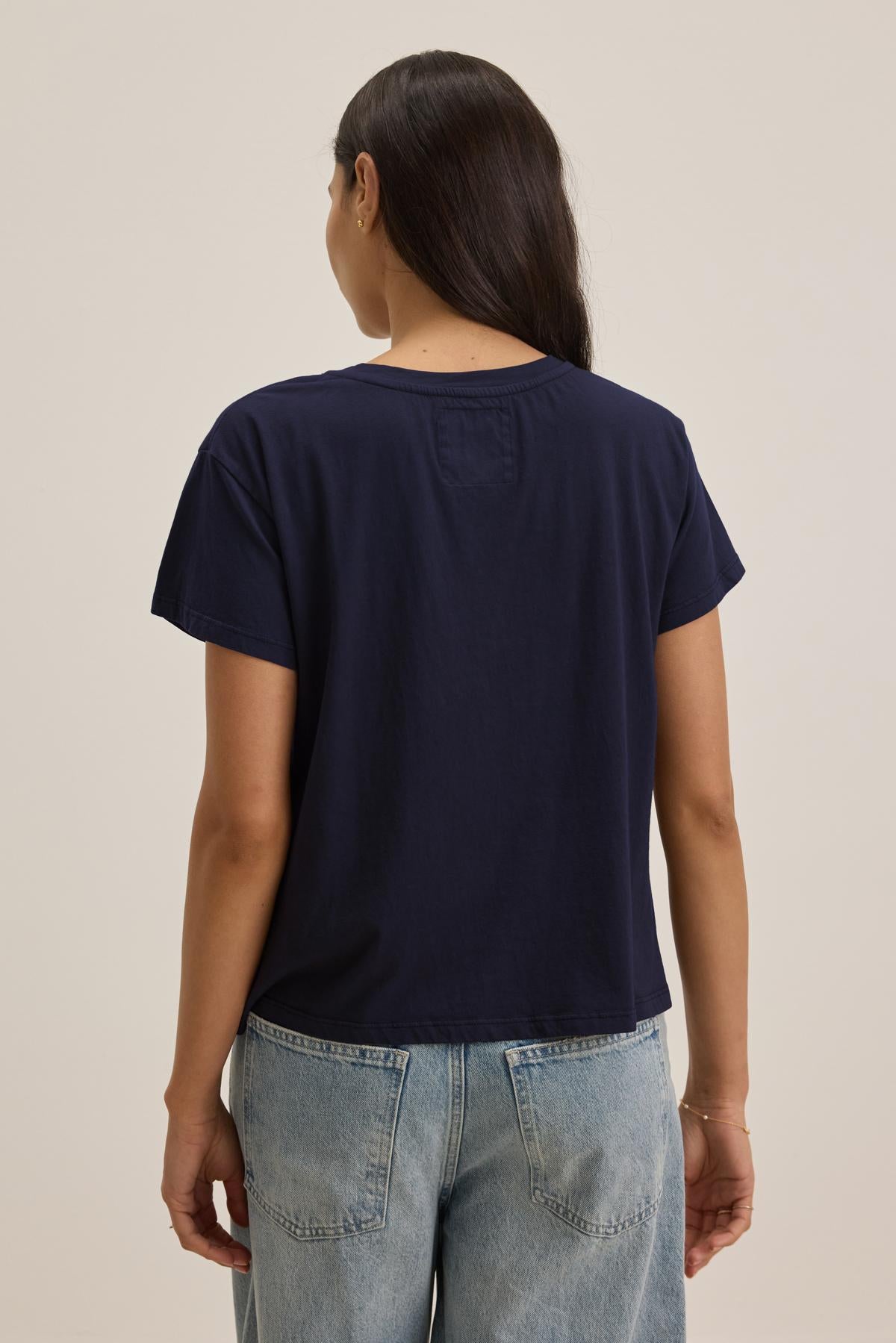 A person with long dark hair wearing a navy blue, relaxed-fit TOPANGA TEE made of organic cotton from Velvet by Jenny Graham and light blue jeans is seen from the back, standing against a plain background.-37817545097409