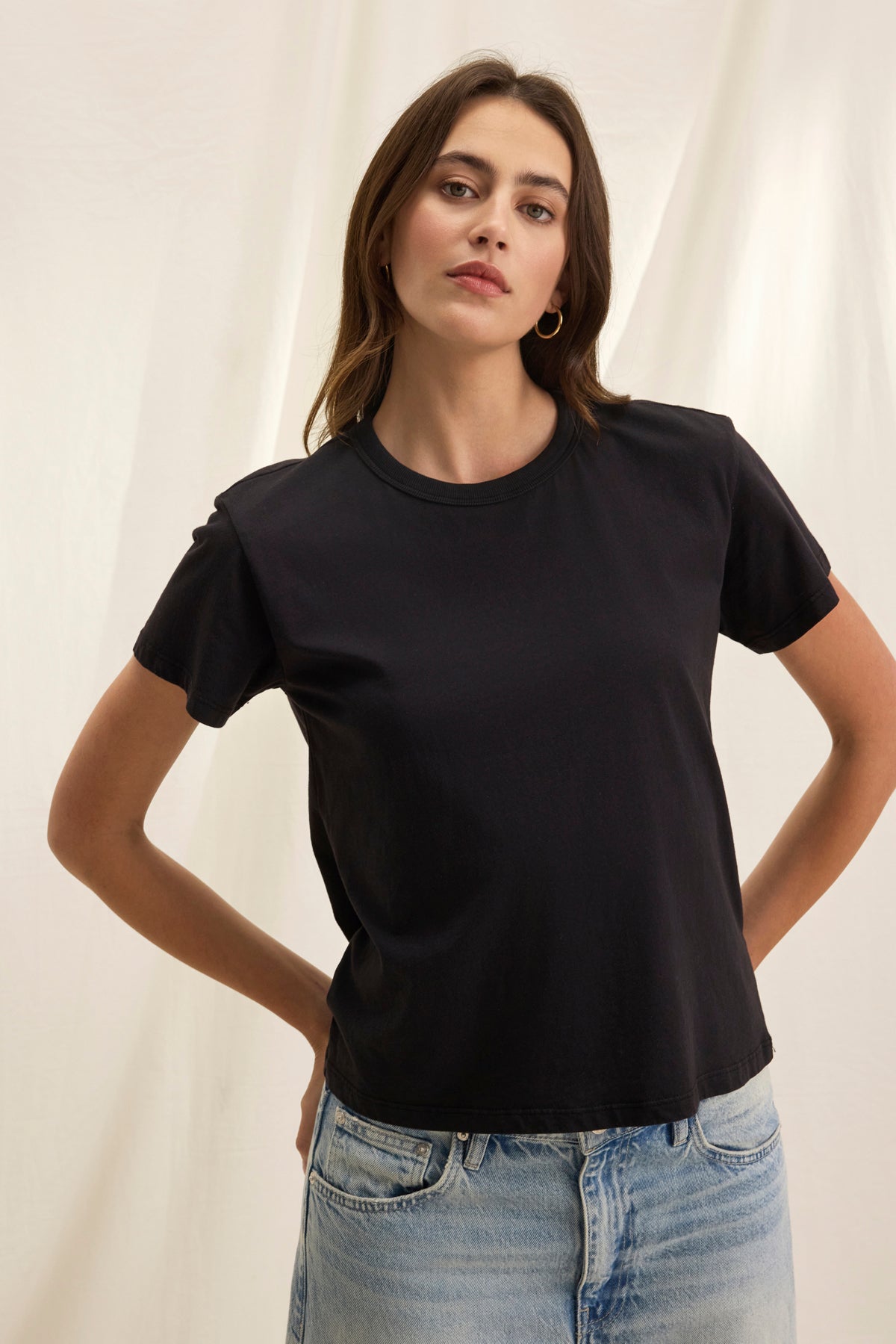   A person in a JAZZ TEE from Velvet by Graham & Spencer, featuring a crew neckline, paired with blue jeans, stands against a light backdrop with hands on hips. 