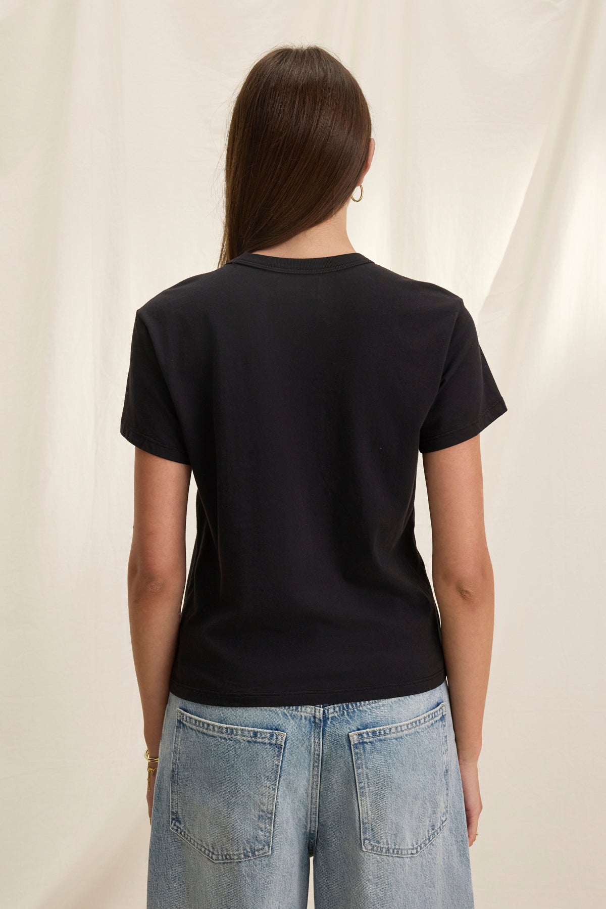   A person with long hair stands back to the camera, wearing a Velvet by Graham & Spencer JAZZ TEE in black. Made from organic cotton jersey, its relaxed boxy fit effortlessly pairs with light blue jeans for a casual yet stylish vibe. 