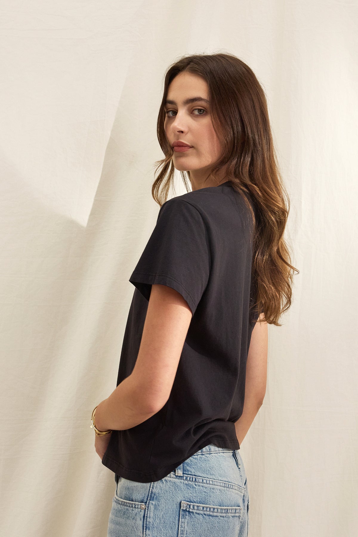   A person with long brown hair in a relaxed silhouette black JAZZ TEE by Velvet by Graham & Spencer, made from organic cotton jersey, pairs it with blue jeans while standing against a light fabric background, looking over their shoulder. 