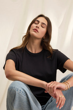 A person sits with eyes closed, surrounded by a white background, wearing Velvet by Graham & Spencer's JAZZ TEE, a black relaxed boxy silhouette t-shirt in organic cotton jersey, paired with jeans.