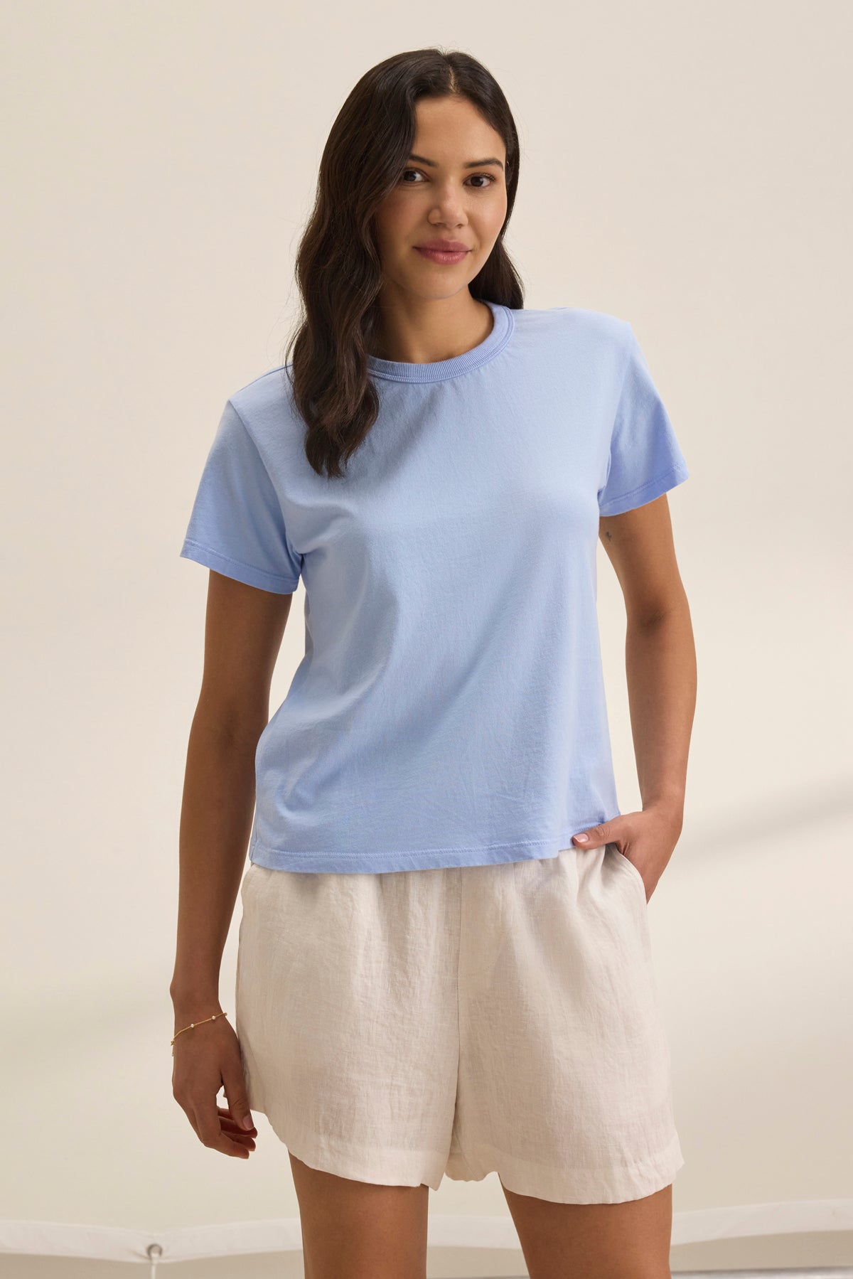   A person with long hair stands against a plain background, wearing the light blue JAZZ TEE by Velvet by Graham & Spencer, paired with white shorts. 