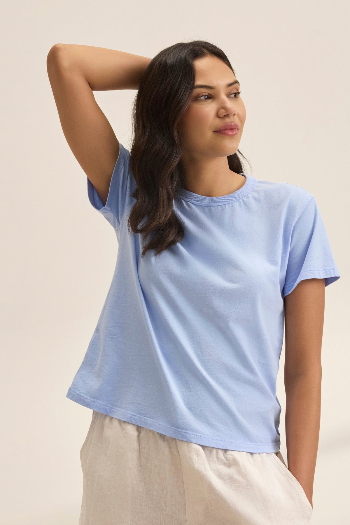   Person wearing a JAZZ TEE by Velvet by Graham & Spencer in light blue organic cotton jersey and relaxed beige pants, posing with one arm raised and looking to the side against a neutral background. 