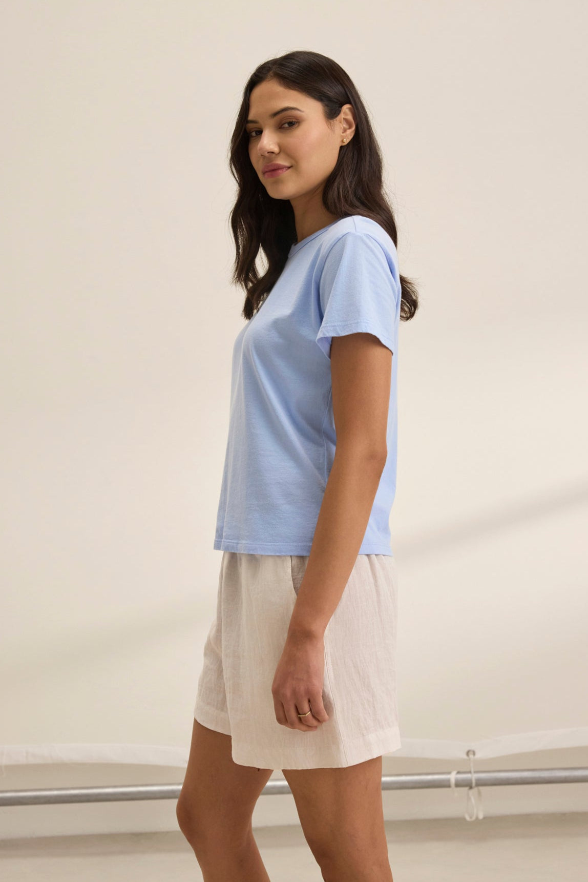   Wearing the JAZZ TEE by Velvet by Graham & Spencer, crafted from light blue organic cotton jersey with a soft crew neckline, a person stands sideways in white shorts in a neutral room, embodying a relaxed silhouette. 