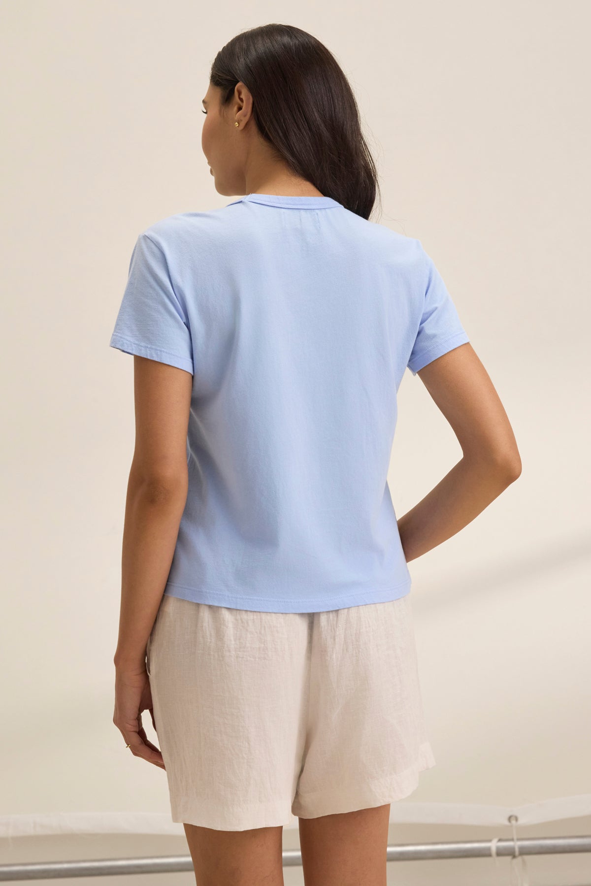   A woman in a light blue JAZZ TEE from Velvet by Graham & Spencer, made of organic cotton jersey, stands with her back to the viewer. The shirt's relaxed boxy silhouette and crew neckline give it an effortlessly casual look when paired with beige shorts. 