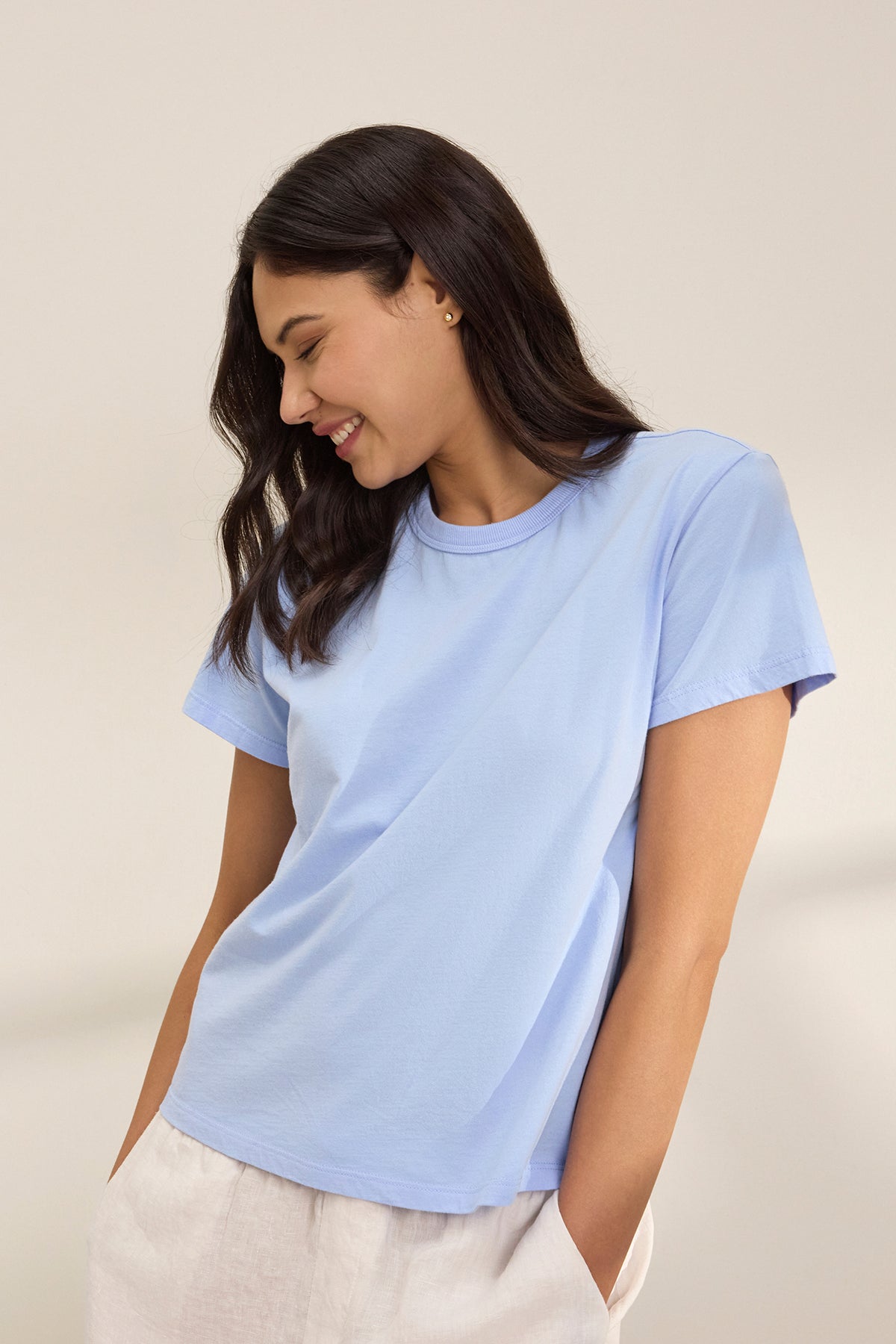   A person in a light blue JAZZ TEE by Velvet by Graham & Spencer and white pants, smiling slightly while looking down against a neutral backdrop. 