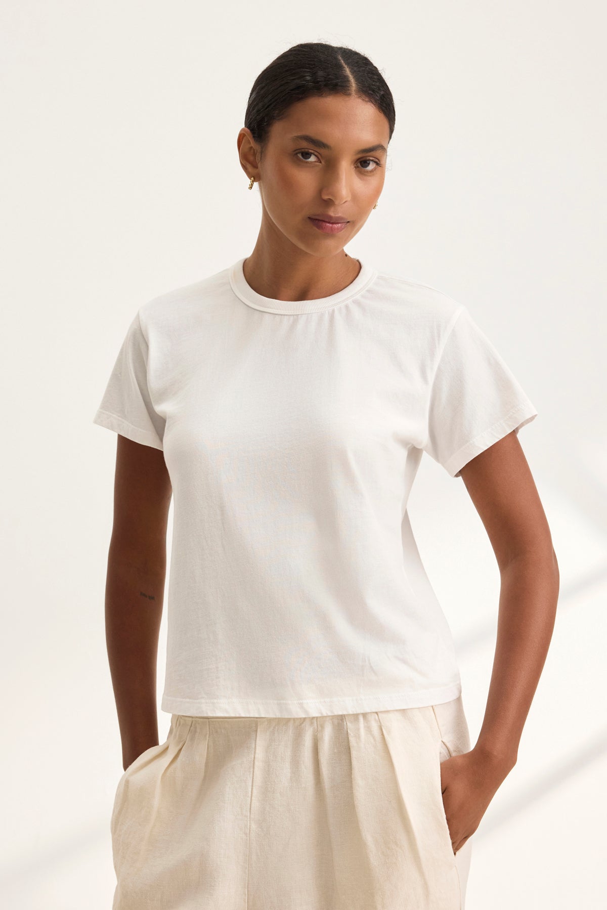   A person wears the JAZZ TEE from Velvet by Graham & Spencer, a white, relaxed boxy silhouette T-shirt in organic cotton jersey with a crew neckline, paired with beige pants. They stand against a plain background with a neutral expression, showcasing the minimalist ensemble. 