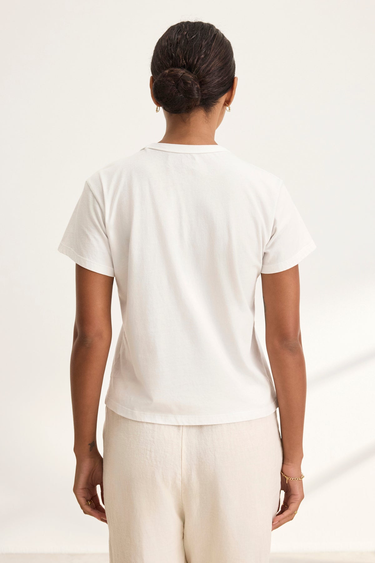 A dark-haired person with a bun is seen from behind wearing a Velvet by Graham & Spencer's JAZZ TEE in white organic cotton jersey, paired with cream pants. The shirt boasts a relaxed boxy silhouette and crew neckline, adding an effortless style against the neutral background.-38819698639041