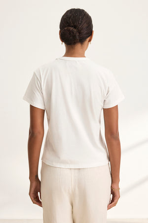 A dark-haired person with a bun is seen from behind wearing a Velvet by Graham & Spencer's JAZZ TEE in white organic cotton jersey, paired with cream pants. The shirt boasts a relaxed boxy silhouette and crew neckline, adding an effortless style against the neutral background.