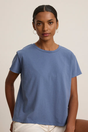 A woman with short, dark hair wears the relaxed-fit TOPANGA TEE in blue by Velvet by Jenny Graham, paired with white pants, standing against a plain, light background.