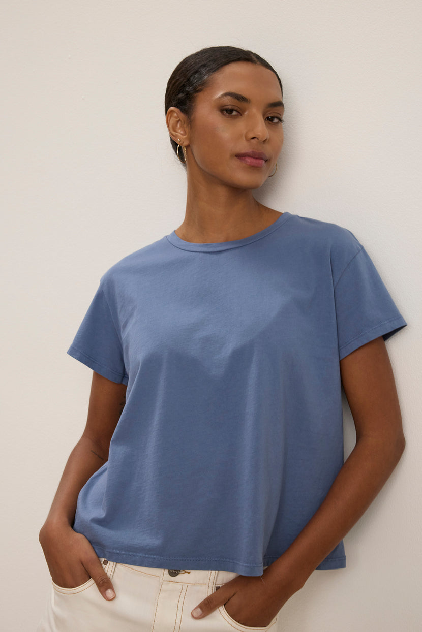 A woman stands against a plain light-colored wall, hands in her pockets, wearing the relaxed-fit TOPANGA TEE by Velvet by Jenny Graham in blue organic cotton and light-colored pants.
