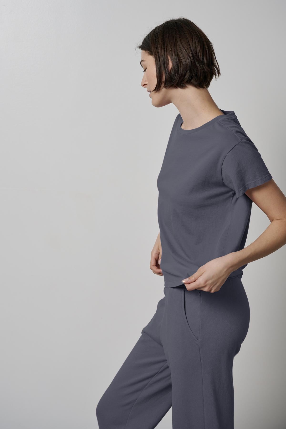 A person with short hair stands against a plain, light-colored wall, wearing a relaxed-fit TOPANGA TEE and pants made from eco-friendly materials by Velvet by Jenny Graham, looking down to their right.-37856362528961