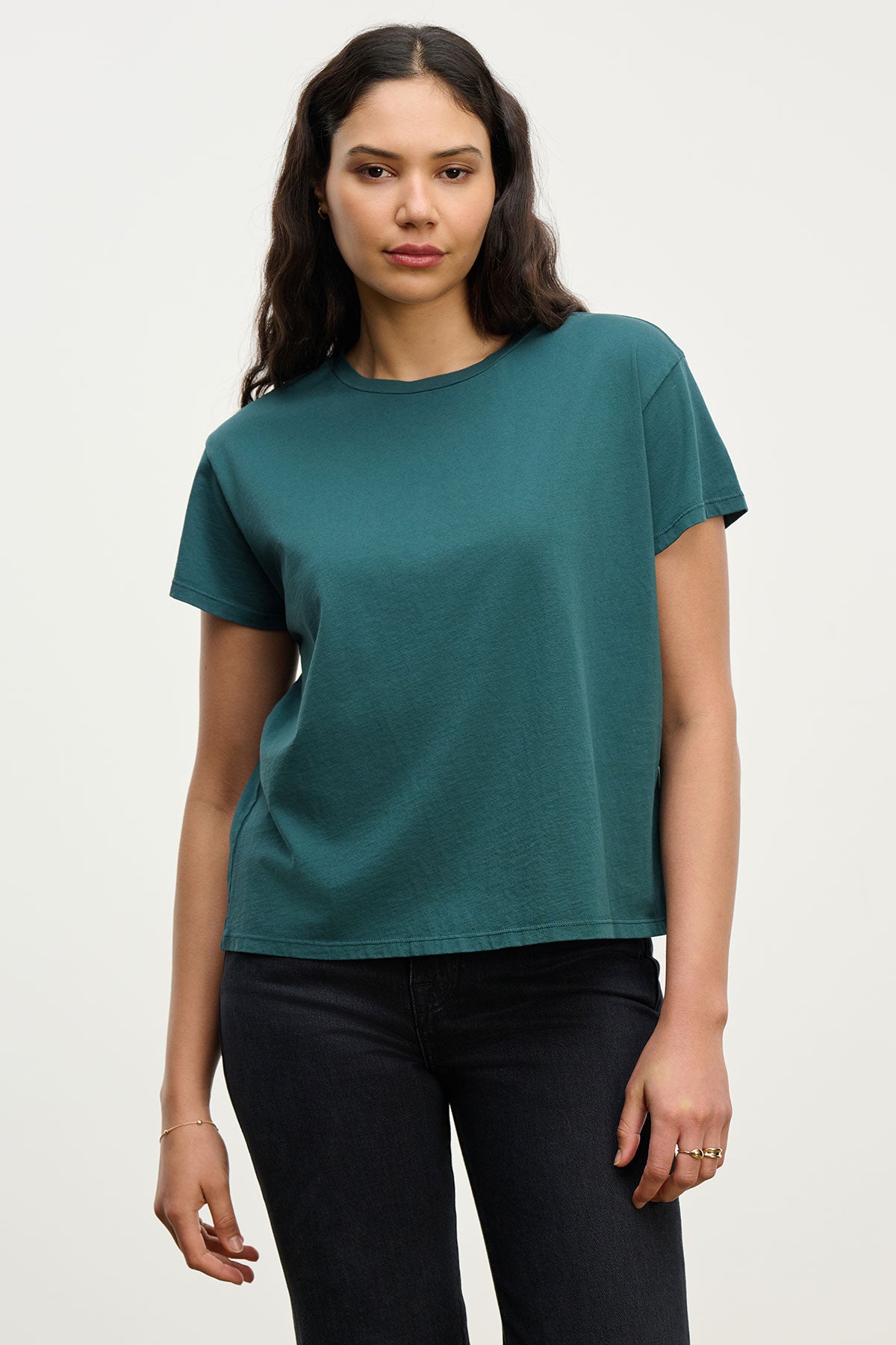   A woman with long dark hair wears the Velvet by Jenny Graham TOPANGA TEE in teal paired with black jeans, standing against a plain light background. 