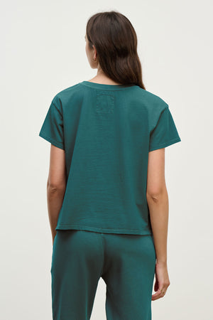 A person with long brown hair is seen from the back, wearing Velvet by Jenny Graham’s TOPANGA TEE and matching teal organic cotton pants—an effortless look that easily takes you from work to workout against a simple light background.