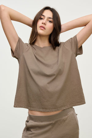 A woman stands with arms raised, wearing the Velvet by Jenny Graham TOPANGA TEE and matching taupe sweatpants—ideal for going from work to workout—against a plain backdrop.