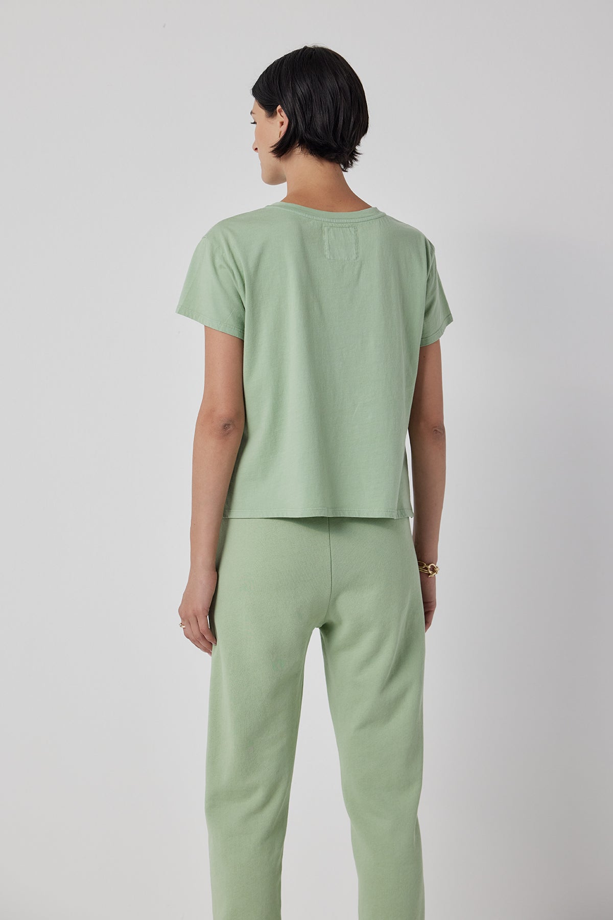   A person with short dark hair is shown from behind, wearing a light green short-sleeve shirt and Velvet by Jenny Graham's WESTLAKE SWEATPANT, which features an elastic drawstring waist. They are standing against a plain white background. 