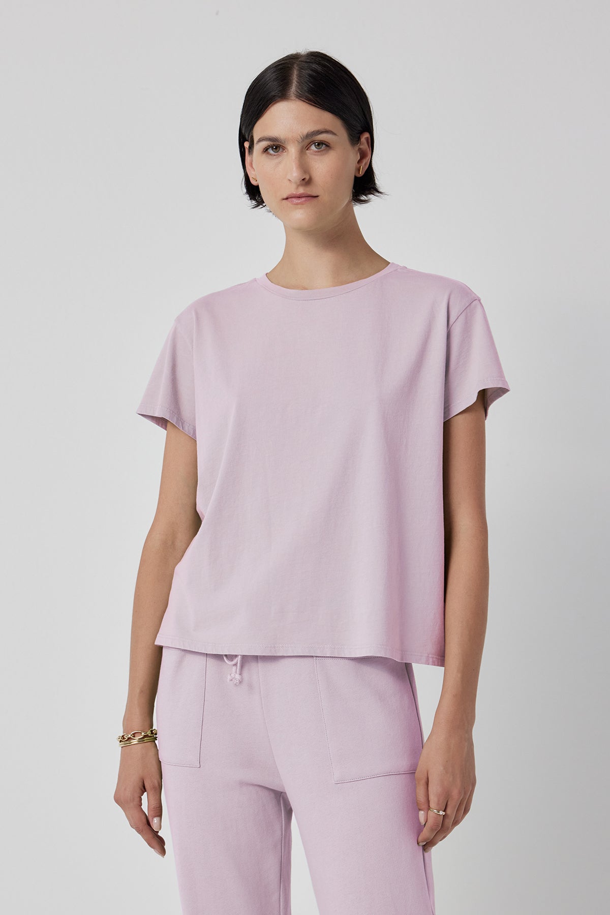 A person stands against a plain background wearing the TOPANGA TEE from Velvet by Jenny Graham, a light pink, relaxed-fit T-shirt paired with matching pants. The outfit, made of organic cotton, complements their short dark hair and neutral expression.-37862282264769