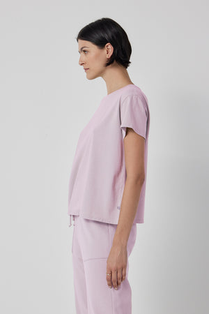 A person stands against a plain background, wearing the short-sleeved, light purple TOPANGA TEE from Velvet by Jenny Graham and matching organic cotton pants. They are looking to the left.